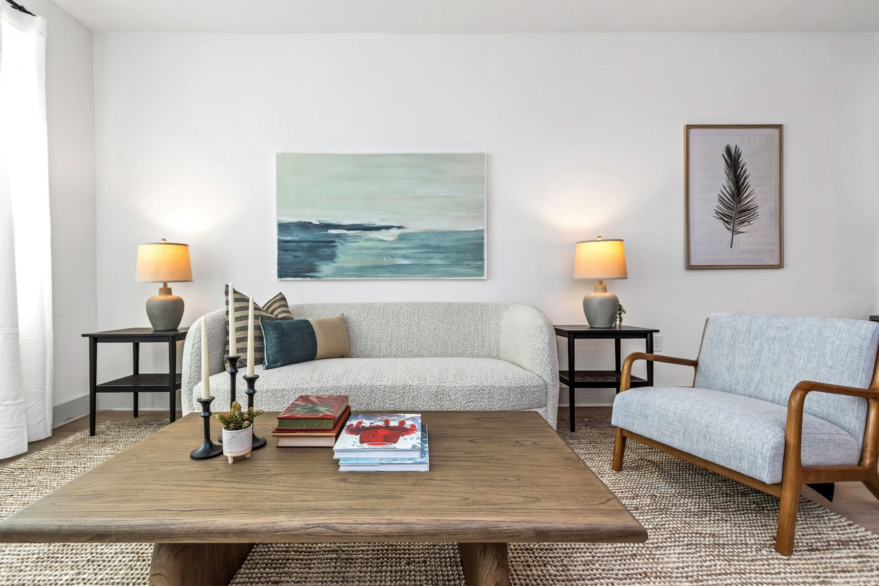 Modern living room with a white sofa, armchair, wooden table, and coastal artwork on the wall.