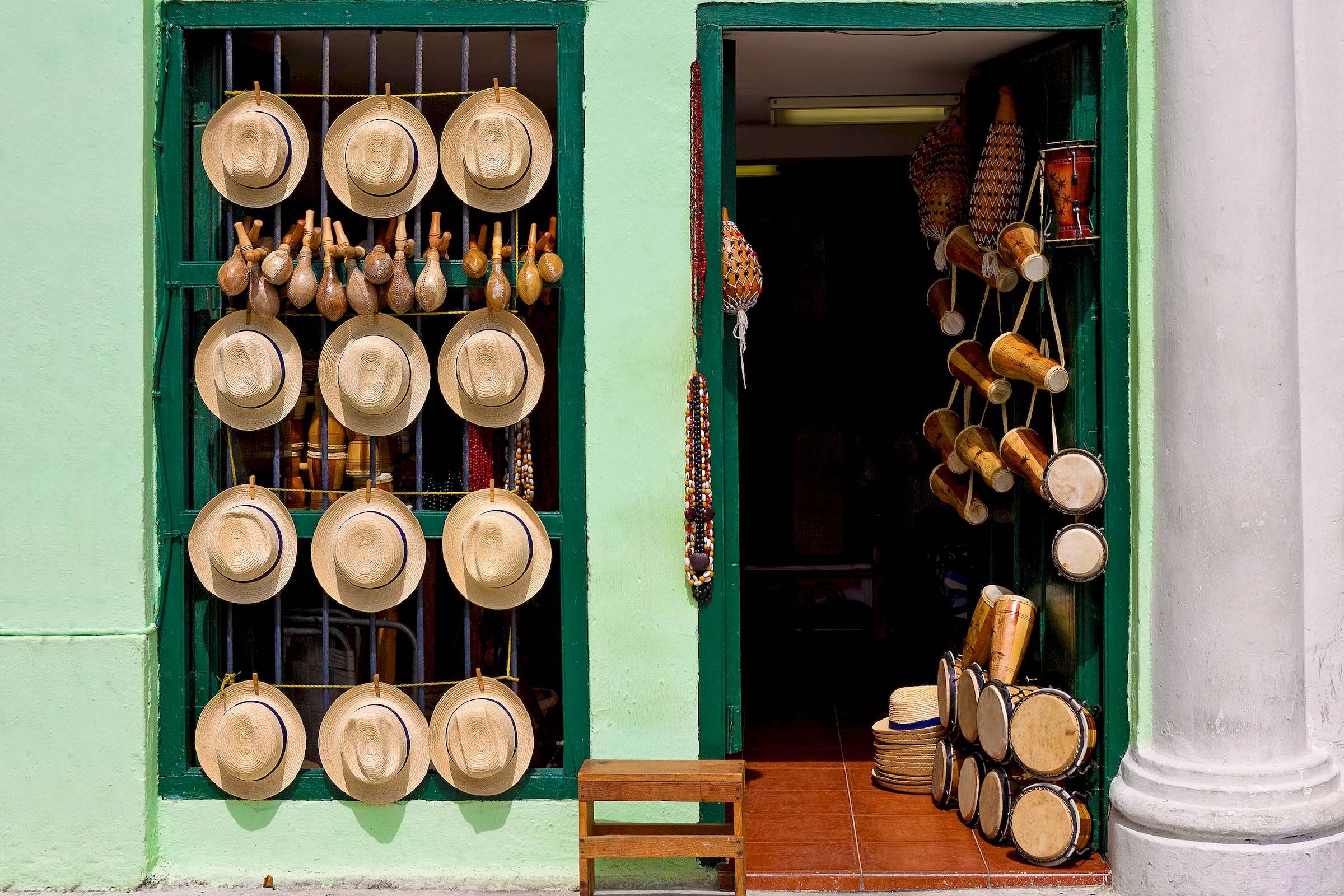 Hats, maracas, and drums displayed outside a green-walled shop with an open door and a wooden stool.