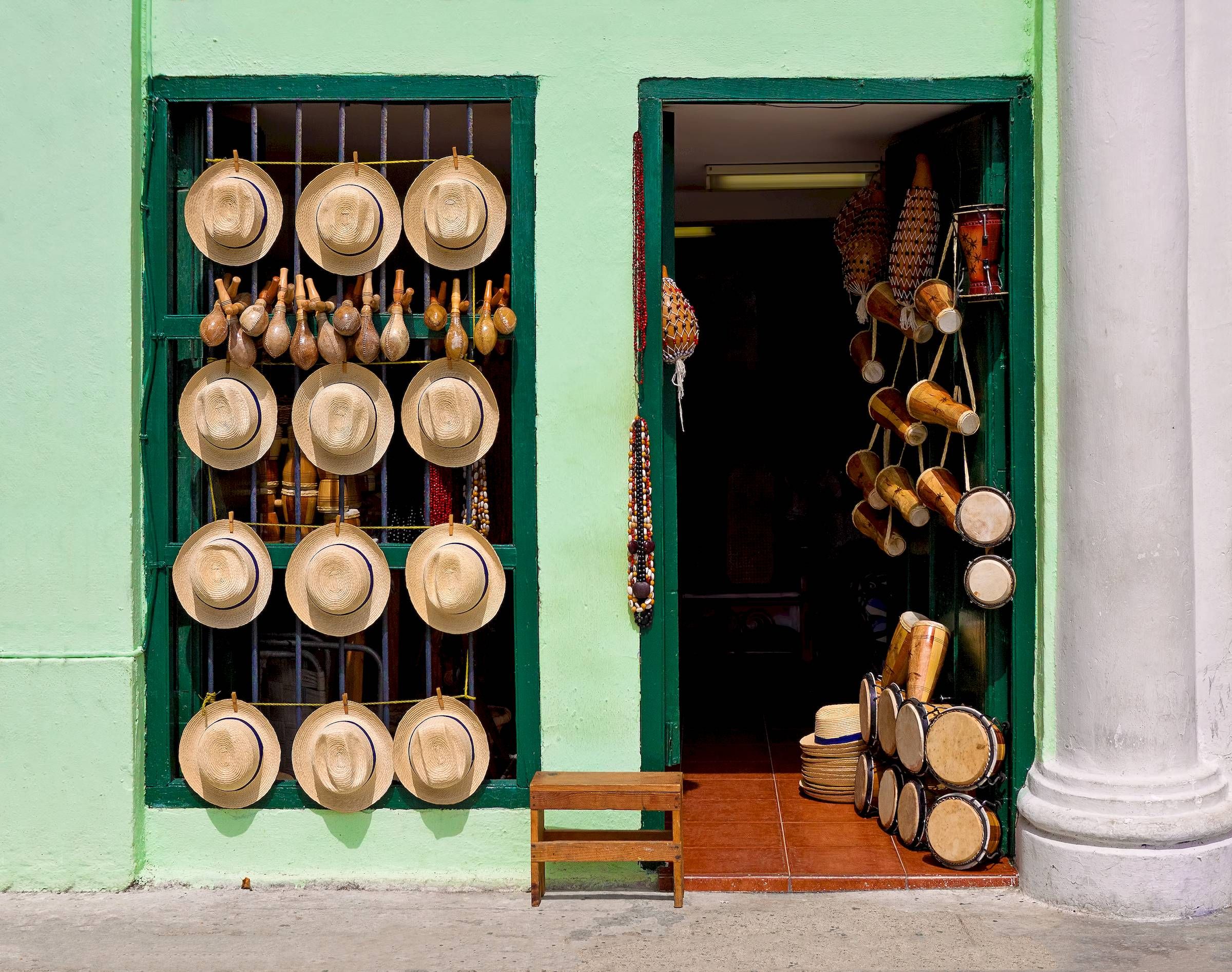 Hats, maracas, and drums displayed outside a green-walled shop with an open door and a wooden stool.