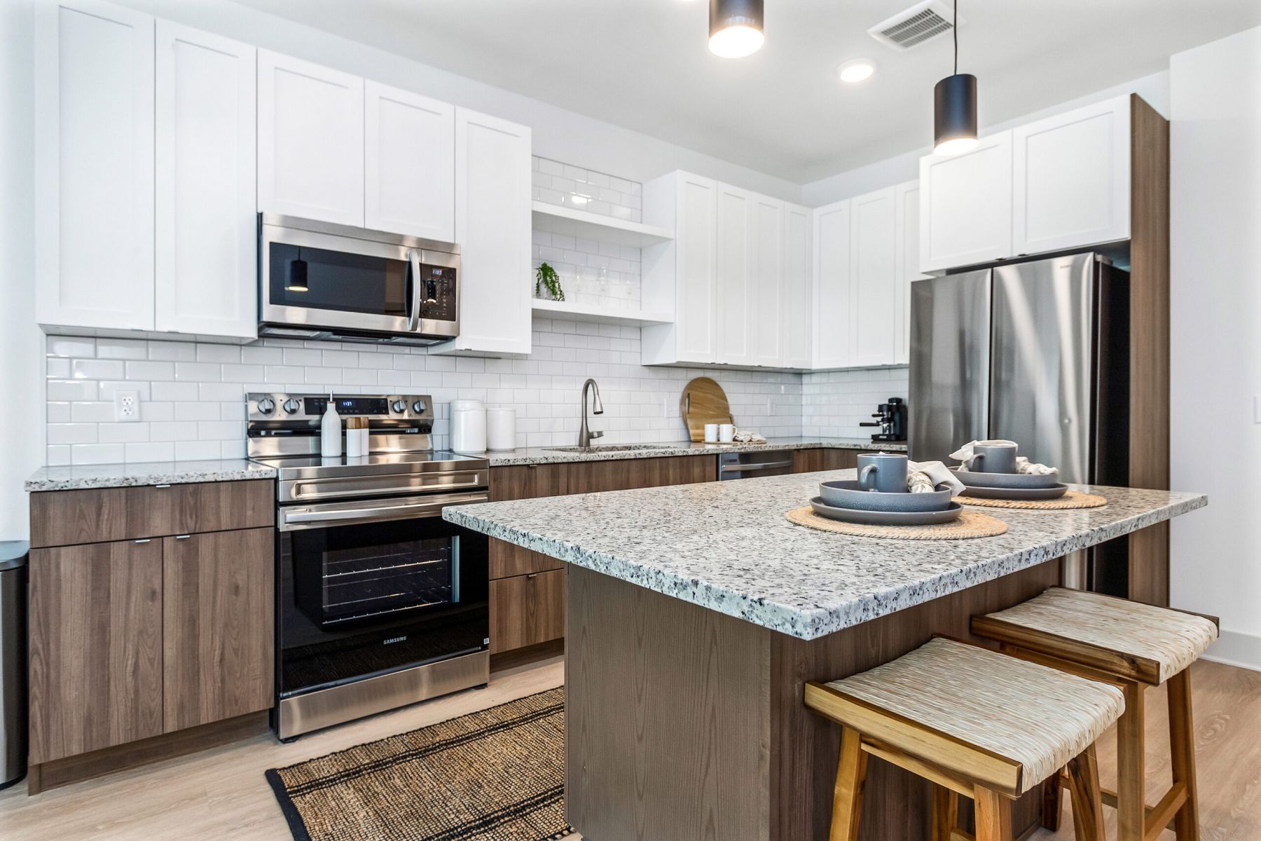 Modern kitchen with stainless steel appliances, granite island, and wooden stools under bright lighting.