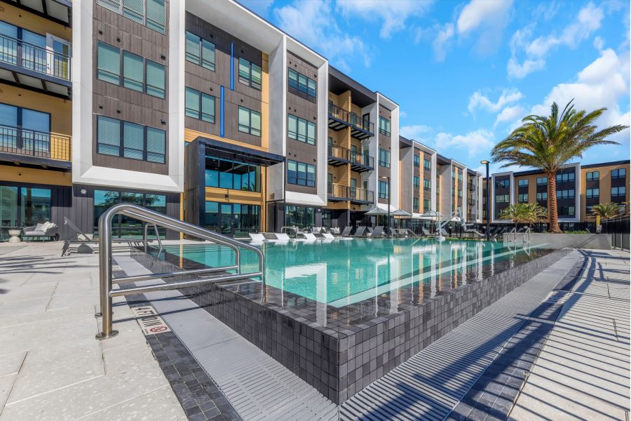 Modern apartment building with balconies overlooking a rectangular outdoor swimming pool under a blue sky.