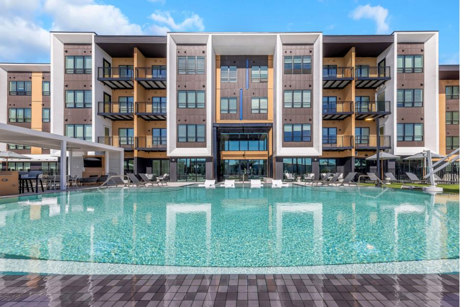 Modern apartment building with balconies overlooking a large, clear swimming pool under a partly cloudy sky.