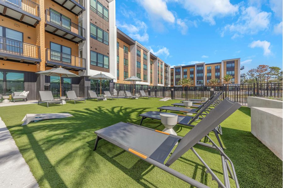 Lounge chairs on a grassy patio outside a modern apartment building, with umbrellas and blue sky above.