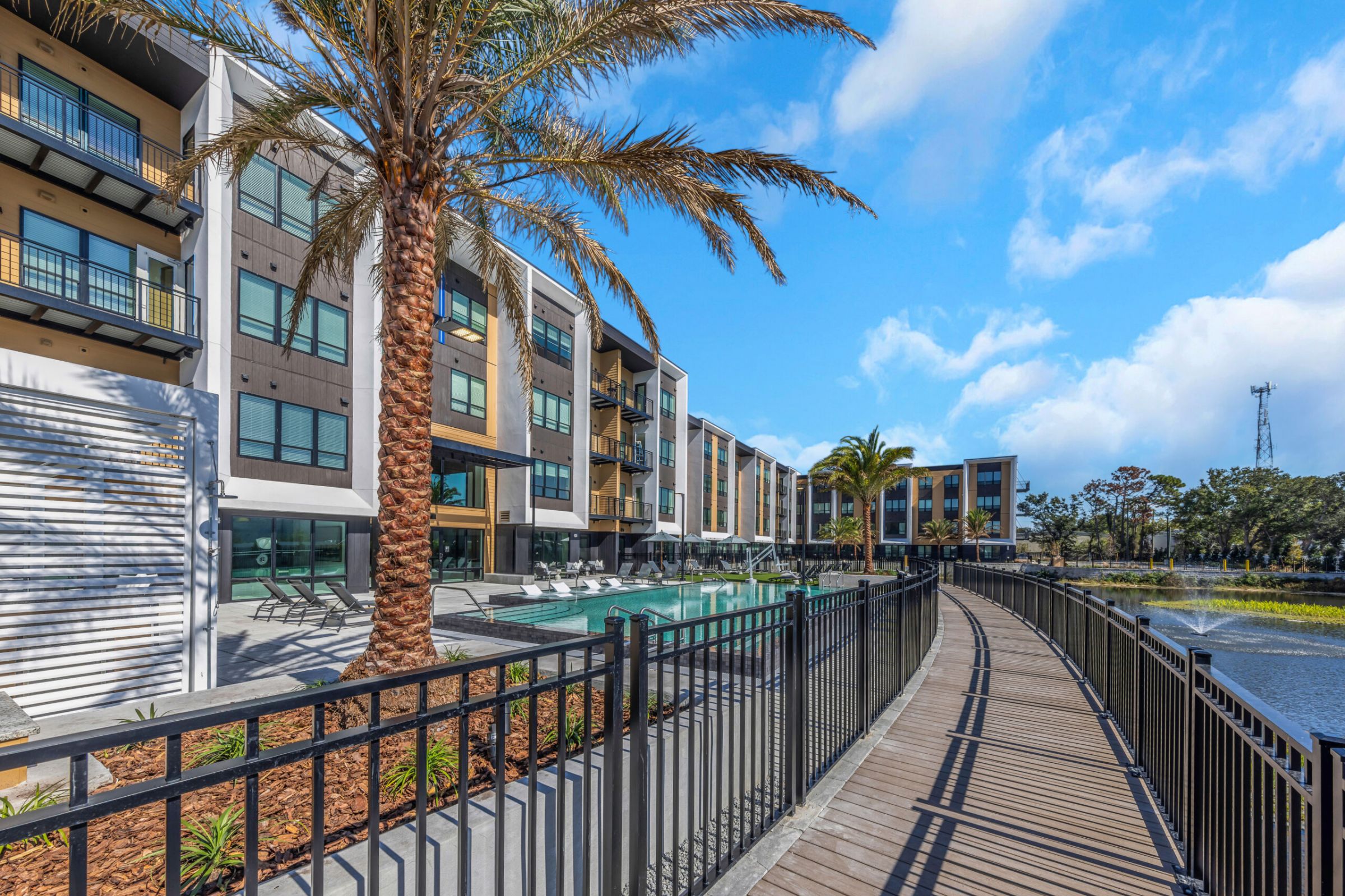 Modern apartment complex with pool, palm trees, and a boardwalk by a pond under a blue sky.
