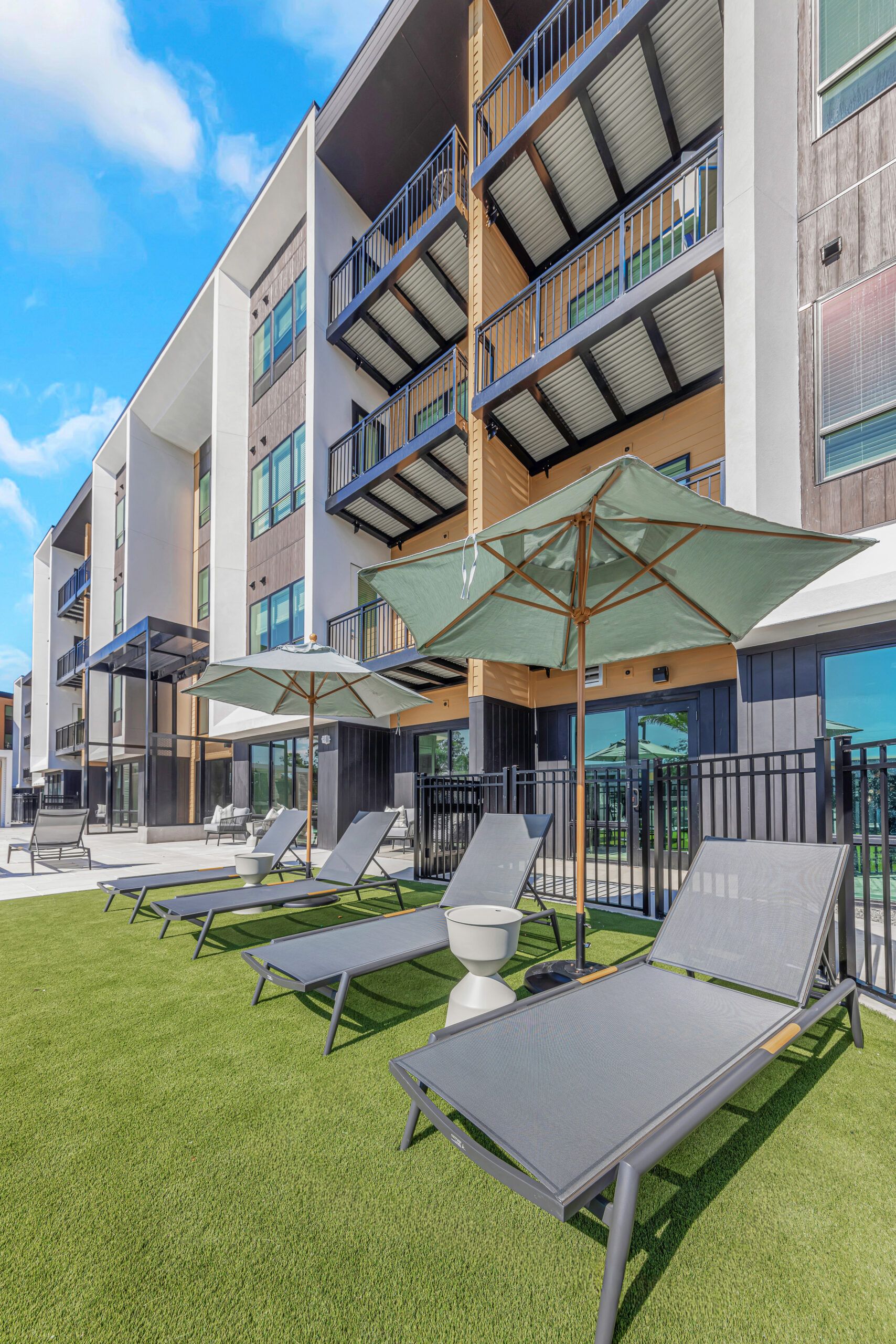 Modern apartment building with lounge chairs and umbrellas on a grassy outdoor area under a bright blue sky.