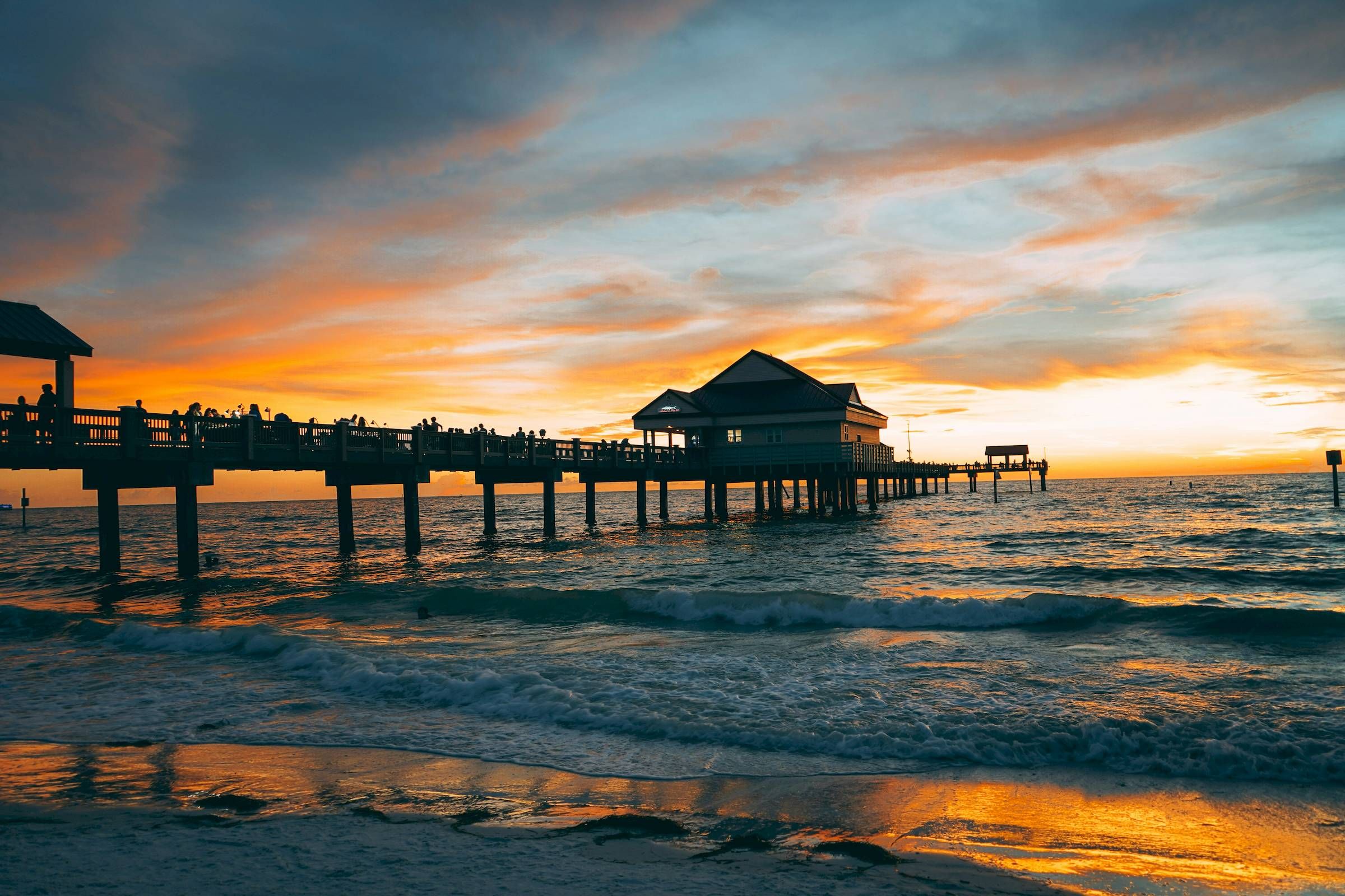 A pier stretching over the ocean at sunset, with vibrant orange and blue skies and gentle waves.