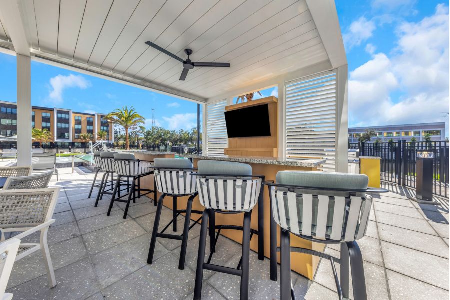 Outdoor bar with striped stools, TV, and ceiling fan under a white pergola on a sunny day.