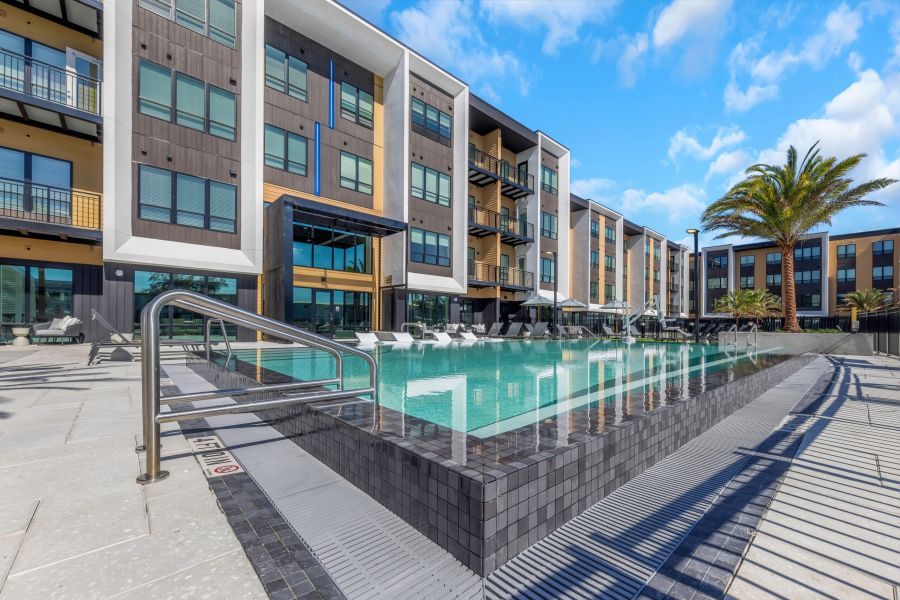 Modern apartment building with balconies overlooking a rectangular outdoor swimming pool under a blue sky.